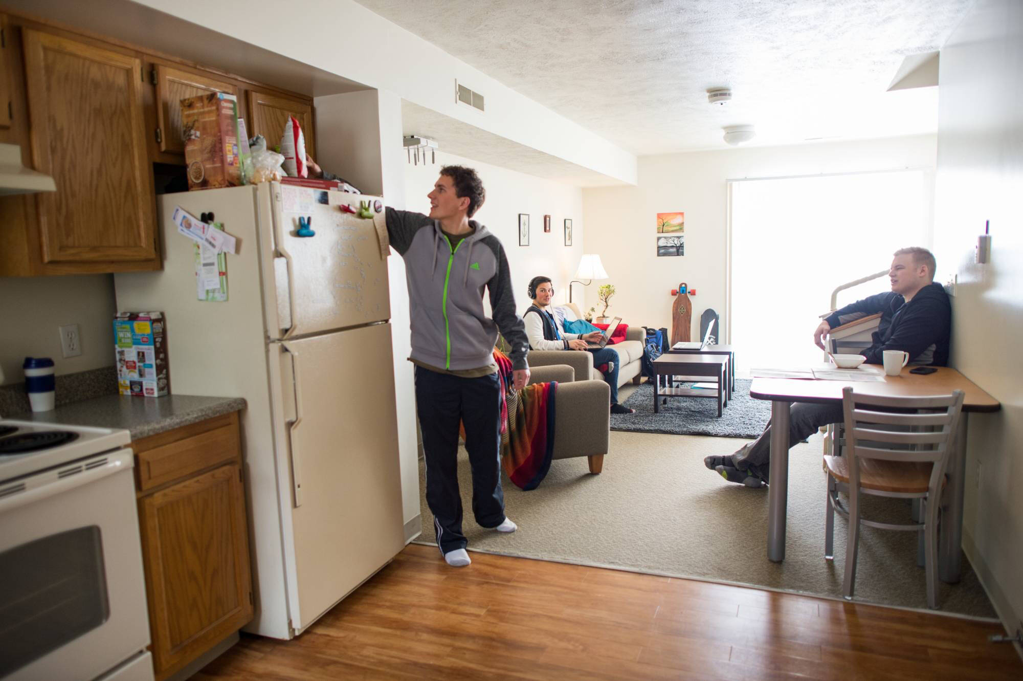 Three people relax in a cozy Lake Village living space. One stands by the fridge, another reclines on a couch, and the third sits at a table with a laptop.